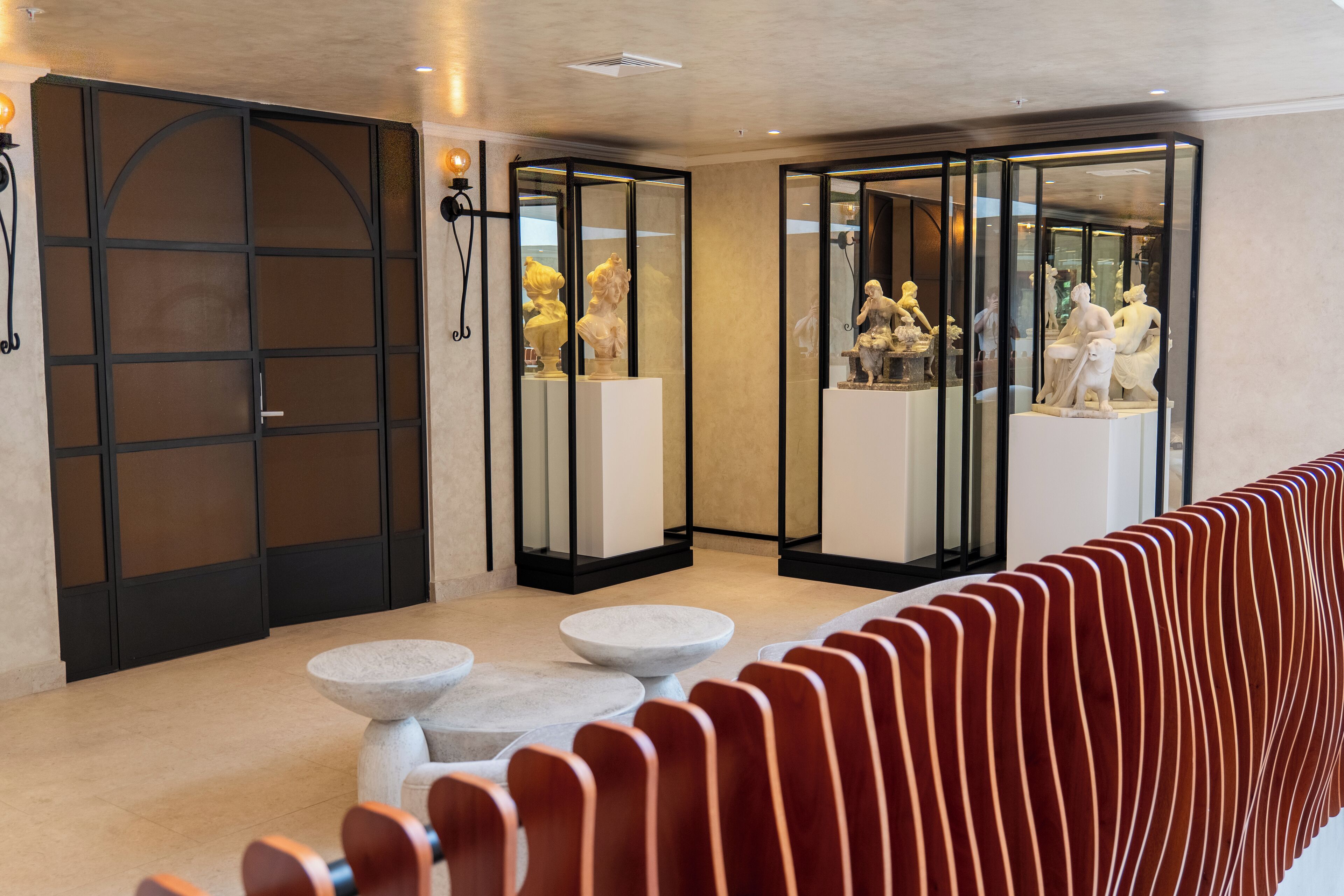 Un moderno vestíbulo de hotel con una hilera larga de sillas rojas, una mesa de piedra y vitrinas de vidrio con esculturas.