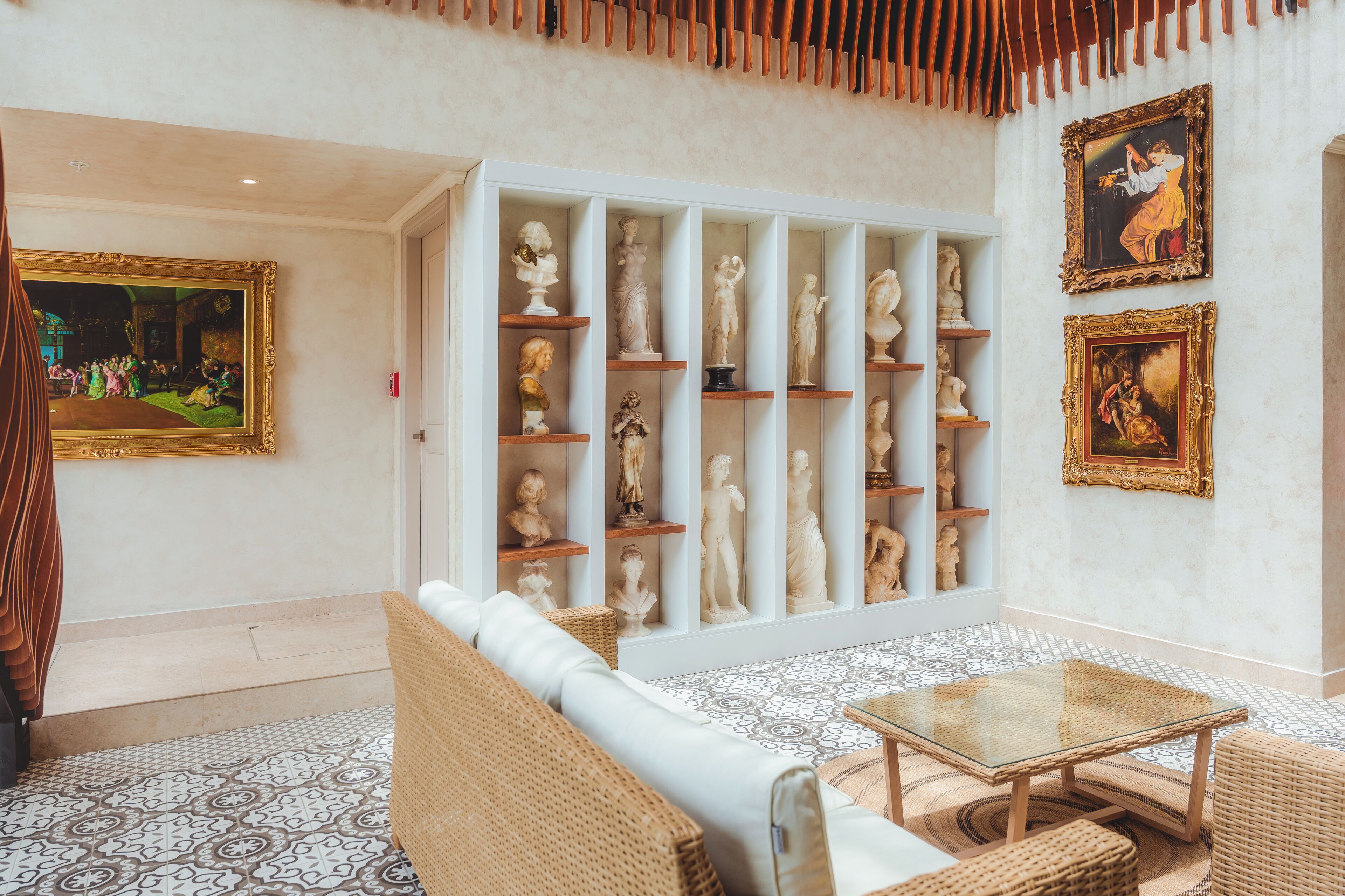A lobby with a white sofa, wicker furniture, a glass coffee table, a white cabinet with sculptures, and framed artwork on the walls.