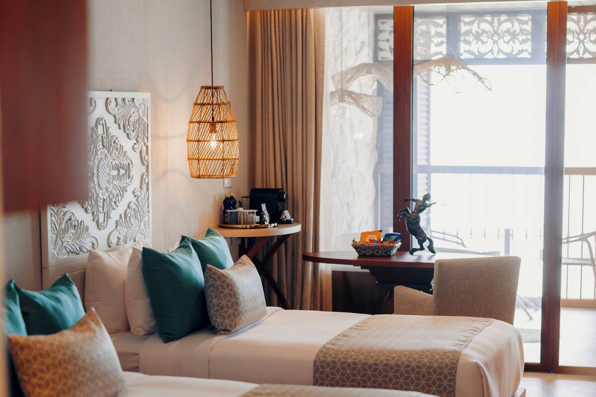 A hotel room with two beds, a desk, a chair, a ceiling fan, and a large window.