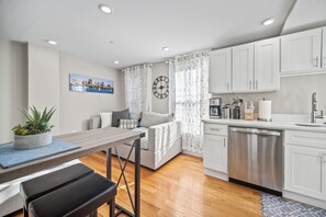 A modern kitchen with white cabinets, a stainless steel dishwasher, and a dining area with a table and bench. - Lux 2nd floor 3brm/1bath (Boston)