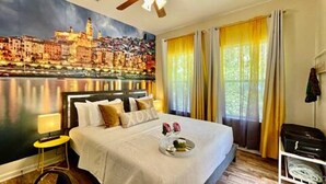 A hotel room with a large bed, a cityscape mural, a ceiling fan, and a window with curtains. - TWO Lake Houses, Sleeps 20, 2 HUGE gAmE rOoMs 407 (Pass Christian)
