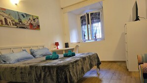 Une chambre à coucher avec un lit, une table recouverte d’une nappe, une fenêtre avec des rideaux, une lampe et un tableau accroché au mur.