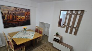 Un coin repas comprenant une table en bois, des chaises, un tableau représentant un paysage urbain, un luminaire au plafond et un miroir muni de crochets.