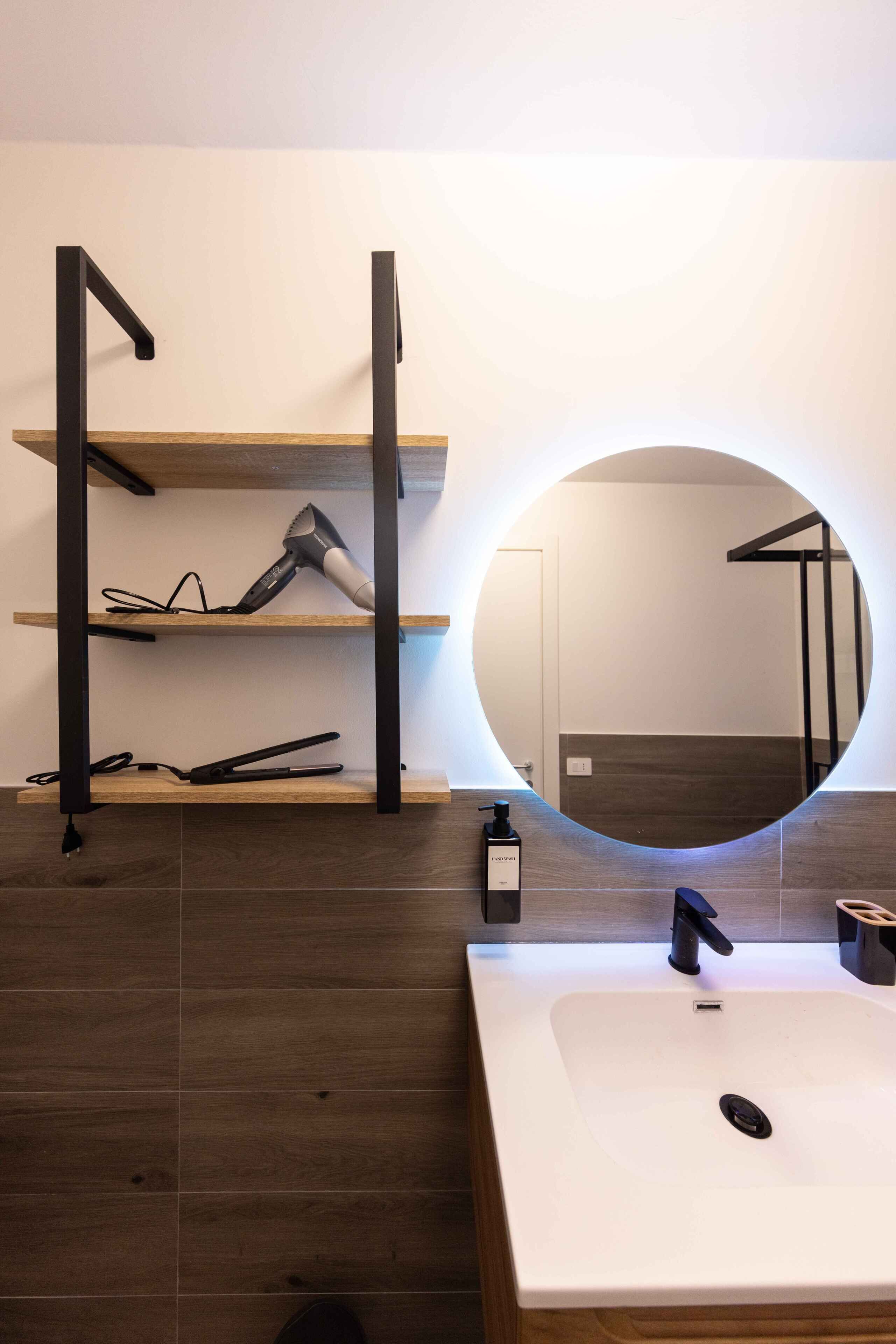 Un bagno moderno con un lavandino bianco, uno specchio rotondo e uno scaffale con un asciugacapelli e una bottiglia.