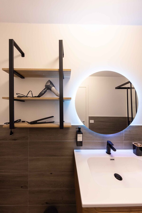 A modern bathroom with a white sink, a round mirror, and a shelf with a hairdryer and a bottle.