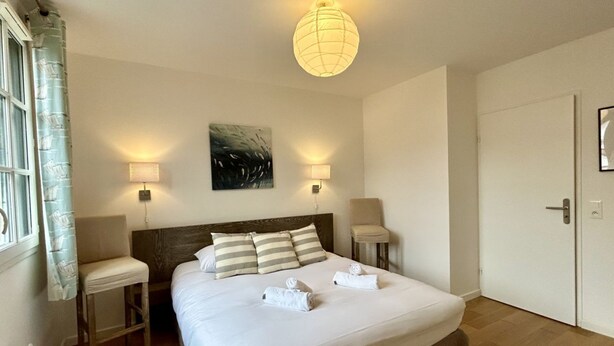A hotel room with a large bed, two bedside tables, a headboard, a painting on the wall, a door, and a window with curtains.