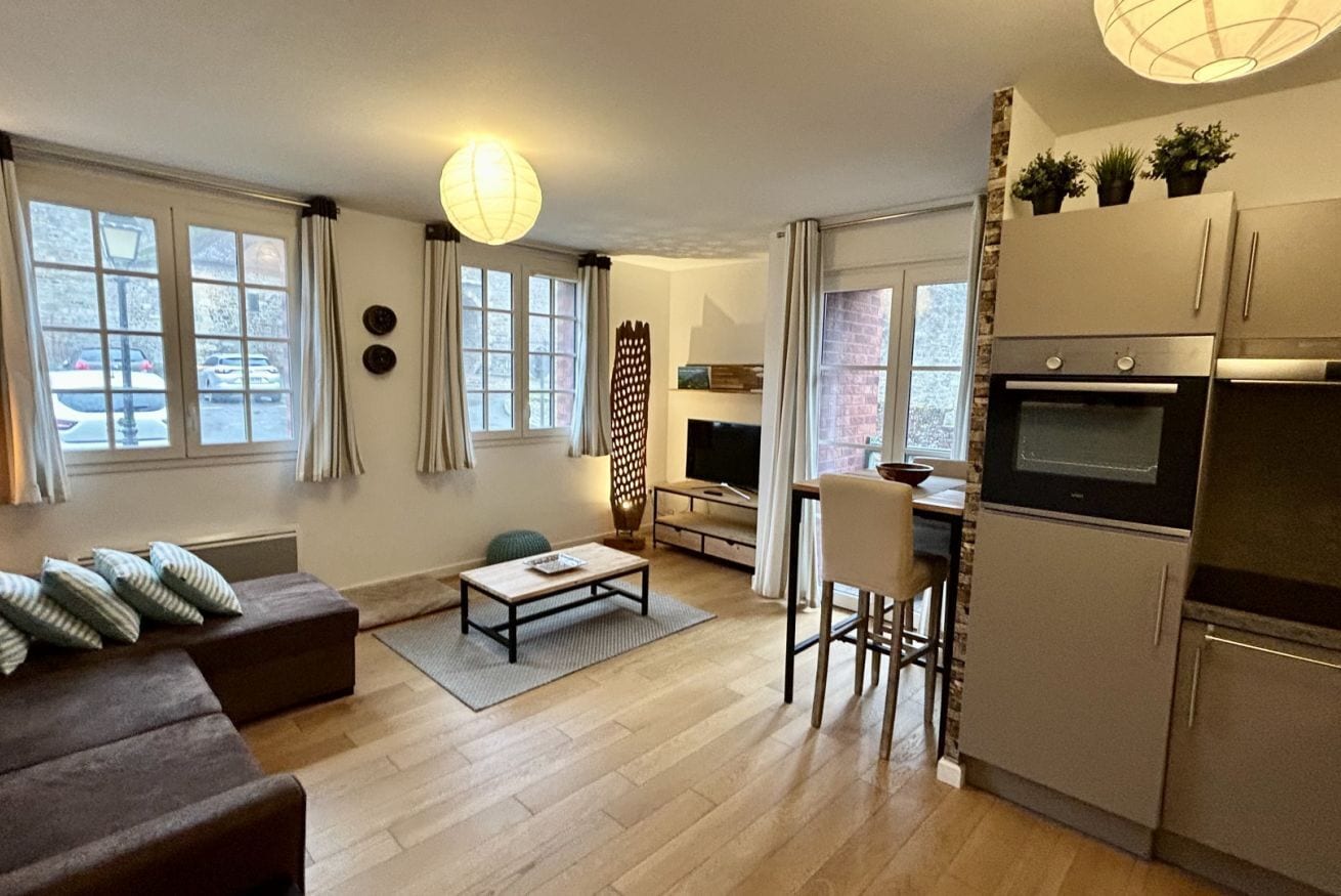 A modern living room with a sofa, a coffee table, and a TV. There is a kitchen with a refrigerator and a microwave.