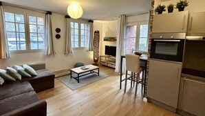 A modern living room with a sofa, a coffee table, and a TV. There is a kitchen with a refrigerator and a microwave.