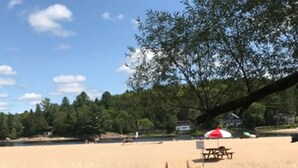 A wooden dock extending into a calm body of water, leading to a sandy beach area with picnic tables and a red umbrella.
