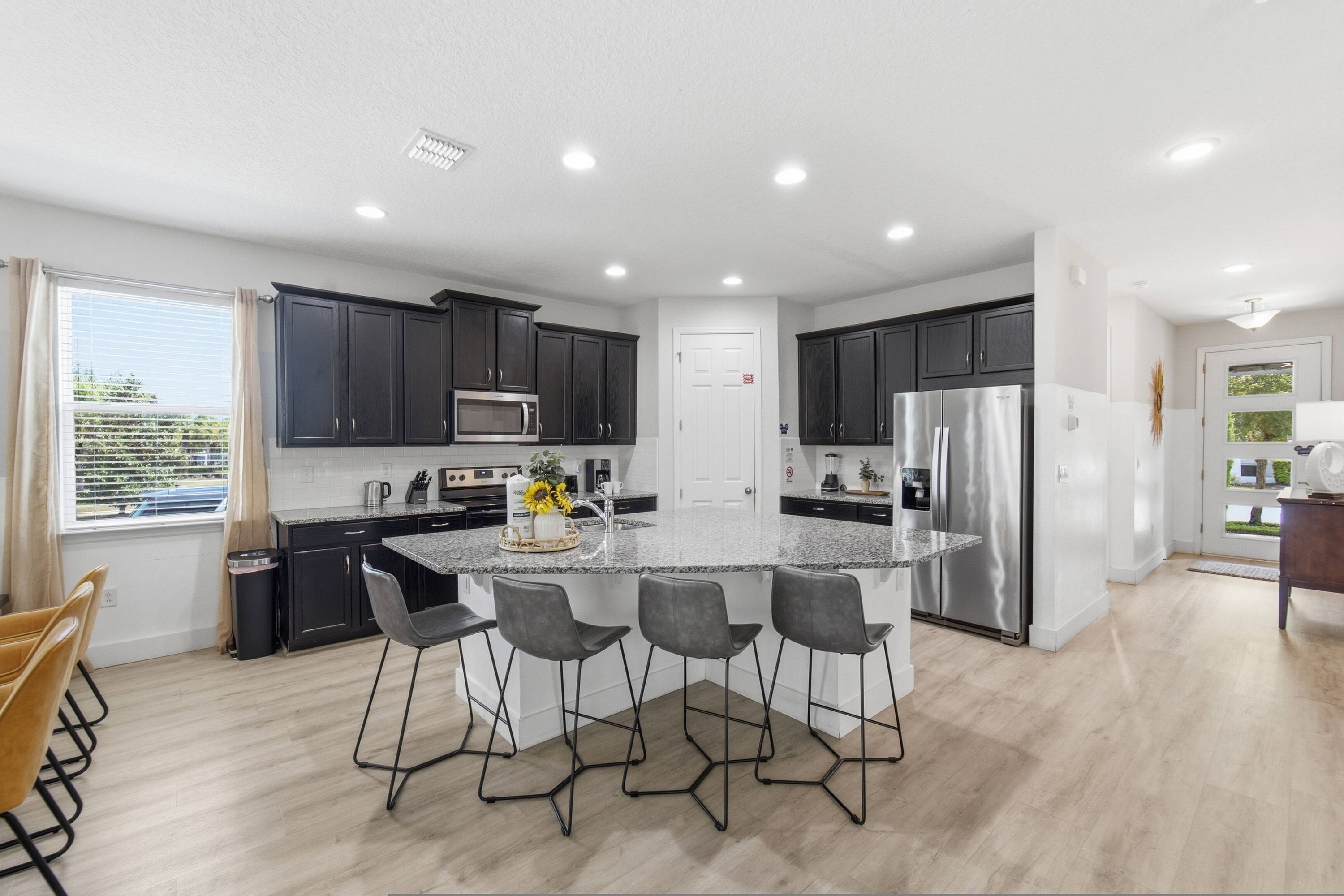 A modern kitchen with dark cabinets, a central island with bar stools, stainless steel appliances, and a view of the outdoors.