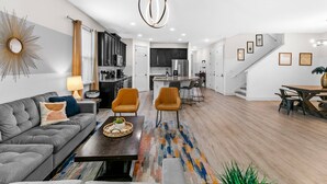 A modern living room with a grey sofa, a coffee table, and orange chairs. There is a kitchen in the background with a stainless steel refrigerator and cabinets. - Spacious 7Bd Next to Clubhouse at Solara Resort (Kissimmee)