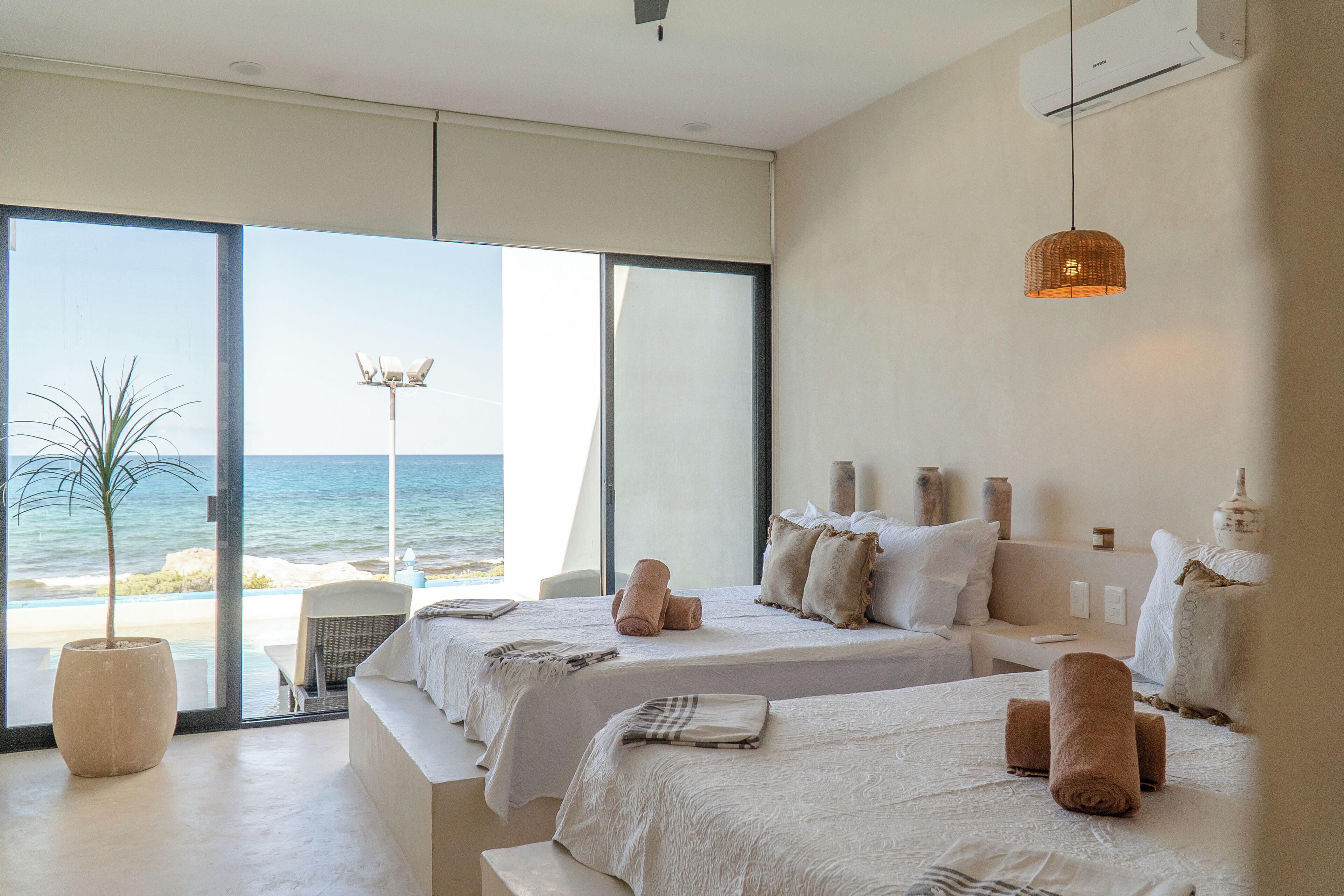 Une chambre d’hôtel avec deux lits, une grande fenêtre donnant sur la plage, une plante en pot et un luminaire suspendu.