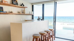 Un espace bar moderne avec vue sur l’océan, des tabourets en bois et des étagères ornées d’objets décoratifs.