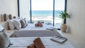 Une chambre avec une grande fenêtre donnant sur la mer, un lit avec du linge de lit blanc, plusieurs oreillers, une plante en pot et une vue sur la plage.