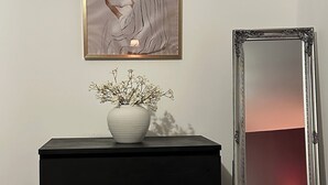 Un aparador negro con un jarrón blanco de flores, un cuadro enmarcado y un espejo que refleja una cama y una lámpara.