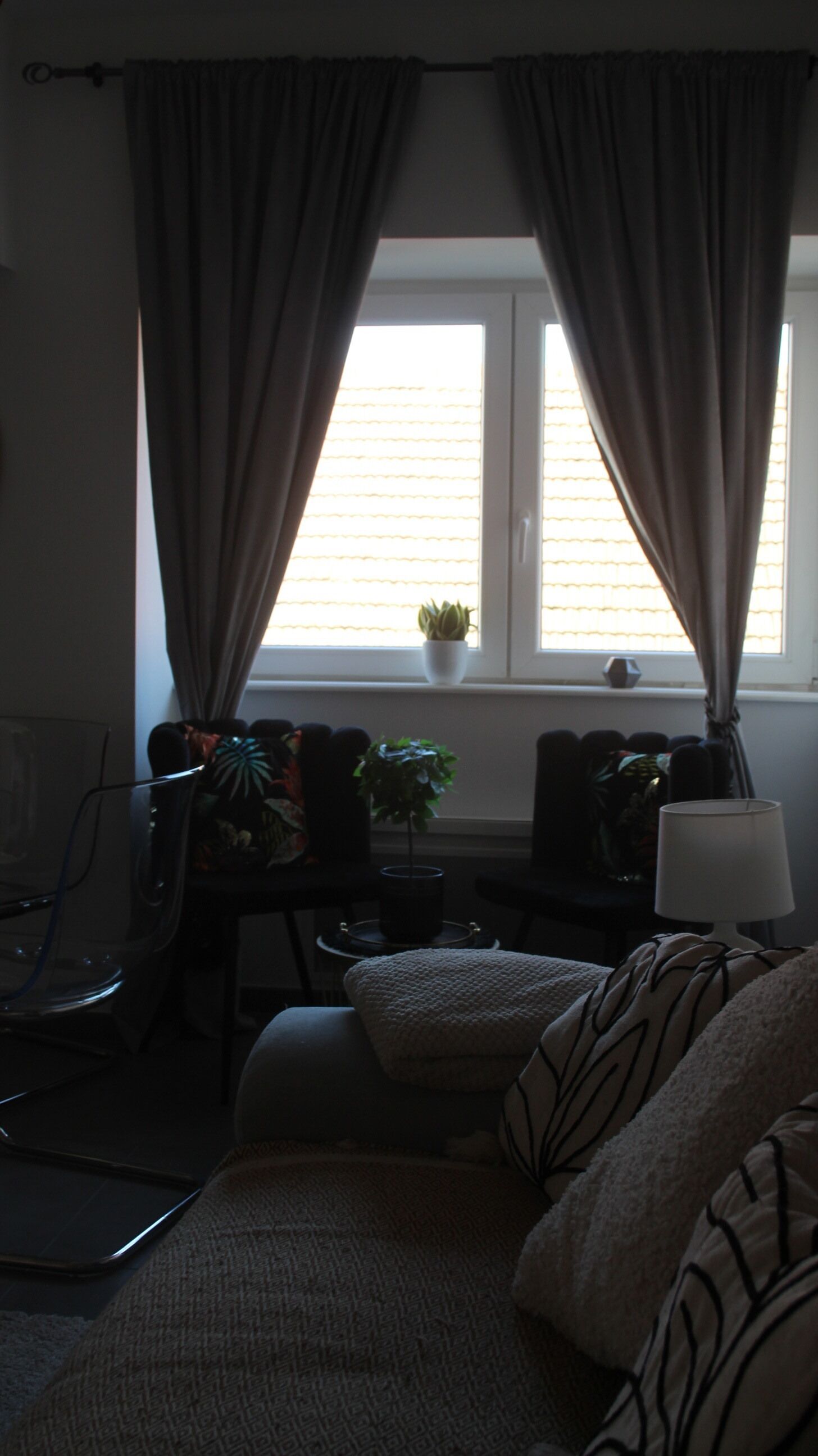 Ein schwach beleuchteter Raum mit einer Couch, einem kleinen Tisch mit einer Lampe und einem Fenster mit Vorhängen.