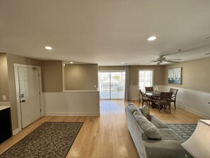A spacious living area with a sofa, dining table, and chairs, a door, and a view through a glass door to the outside.