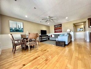 A spacious living room with a wooden floor, a dining area with wicker chairs, a grey sofa, and a flat-screen TV. - Tybee Retreat Whole Building (Tybee Island)