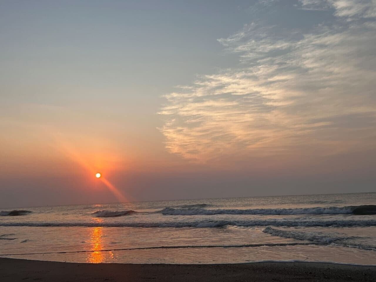 A beach scene with the sun setting over the ocean, waves approaching the shore, and a clear sky.