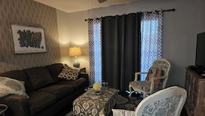 A living room with a brown sofa, patterned ottoman, and a white armchair with blue floral upholstery. A lamp and a framed picture adorn the wall.
