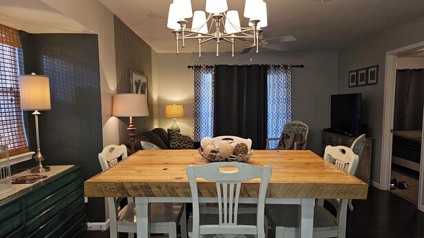 A dining area with a wooden table, white chairs, a chandelier, and a television in the background.