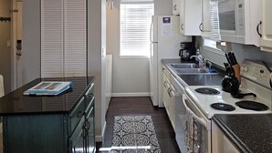 A compact kitchen with white cabinets, a stove, microwave, and a refrigerator. There is a window with blinds, a clock, and a door with a lock.