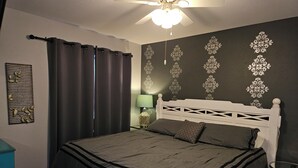 A bedroom with a bed, a ceiling fan, a nightstand, a lamp, and a decorative wall panel.