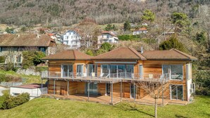 Une maison moderne à deux étages, construite en bois et dotée d’un balcon, qui s’intègre dans un paysage vallonné, entourée d’autres immeubles résidentiels.