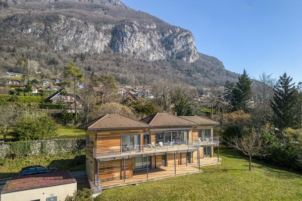 A modern two-story wooden house with a balcony, set against a backdrop of a mountain and a residential area.