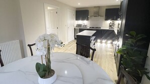 Une cuisine moderne avec des meubles sombres et un îlot central, un coin repas avec une table blanche et un arrangement floral d’orchidées, et une vue sur l’espace de vie adjacent.