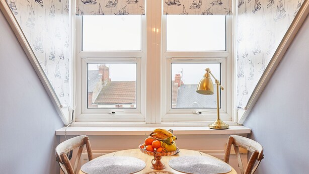 Un coin repas avec une table dressée pour deux personnes, un vase rempli de fruits et une lampe posée sur un rebord de fenêtre.