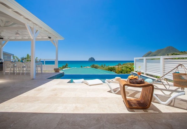 Relax by the ocean with a serene view of a distant island, a pool, and a patio area with lounge chairs and dining space.