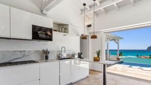 A modern kitchen with white cabinetry, a marble backsplash, and a view of the ocean through an open patio. - Cézembre (Le Diamant)