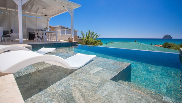 A infinity pool with clear blue water, white lounge chairs, and a covered seating area with a view of the ocean and a distant island. - Cézembre (Le Diamant)