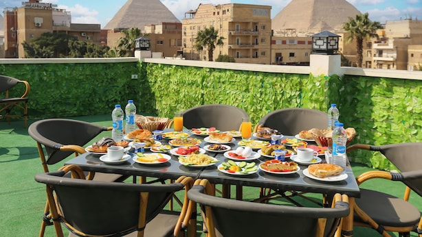 A rooftop dining table with various dishes, surrounded by chairs, overlooking a cityscape with pyramids in the background. - Nefertiti pyramids (Giza)