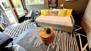 A modern bedroom with a bed, a small table, and a chair. There is a window and an air conditioning unit on the wall. - Charming, bungalow + hot tub in NODA district of Charlotte. 10 min f/uptown. (Charlotte)