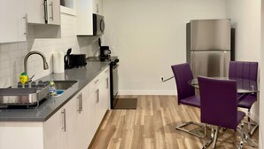 Una cucina compatta con mobili bianchi, un frigorifero in acciaio inossidabile, un lavello e un piccolo angolo ristoro con sedie viola.