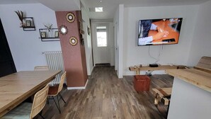 A modern living space with a wooden dining table, chairs, a TV mounted on the wall, and a hallway leading to a door. - Renovated and elegant Ledoux Land house near Disney and 45 min from Paris (Bailly-Romainvilliers)