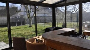 A conservatory with a curved wooden table and chairs, large glass windows, and a view of a backyard with a lawn and a shed. - Renovated and elegant Ledoux Land house near Disney and 45 min from Paris (Bailly-Romainvilliers)