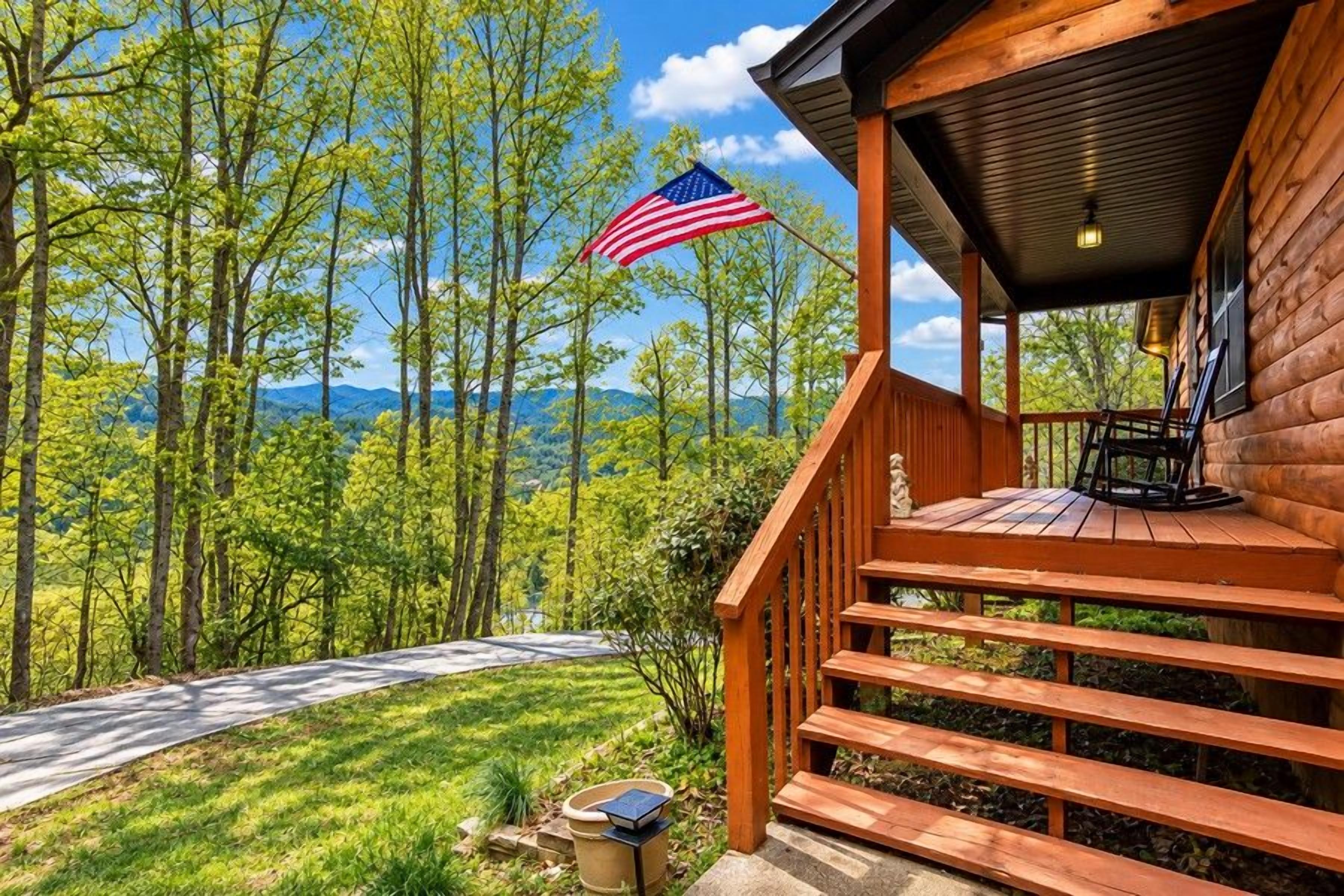 A wooden cabin with a porch, a flag, and a view of a forested area.