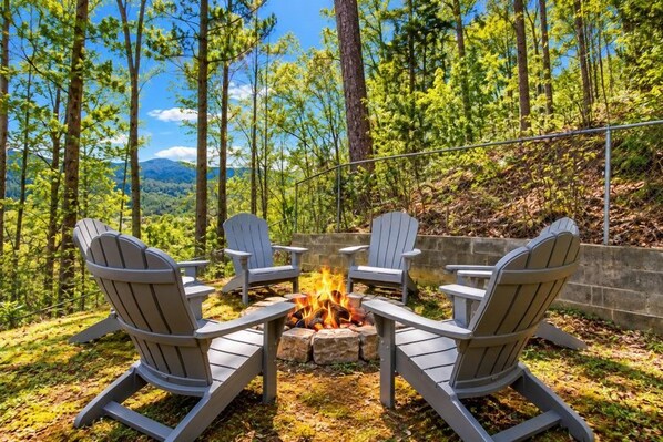 An outdoor seating area with Adirondack chairs around a fire pit, surrounded by a forested landscape.
