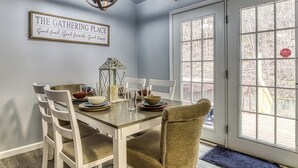 Un comedor con una mesa dispuesta para cuatro, sillas blancas, una araña y un letrero que dice "THE GATHERING PLACE".