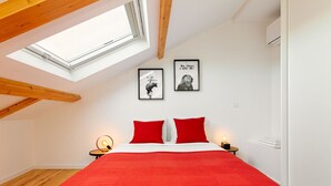 Un dormitorio con tragaluz, una cama con ropa de cama roja, dos cuadros en la pared y una mesita de noche con una lámpara.