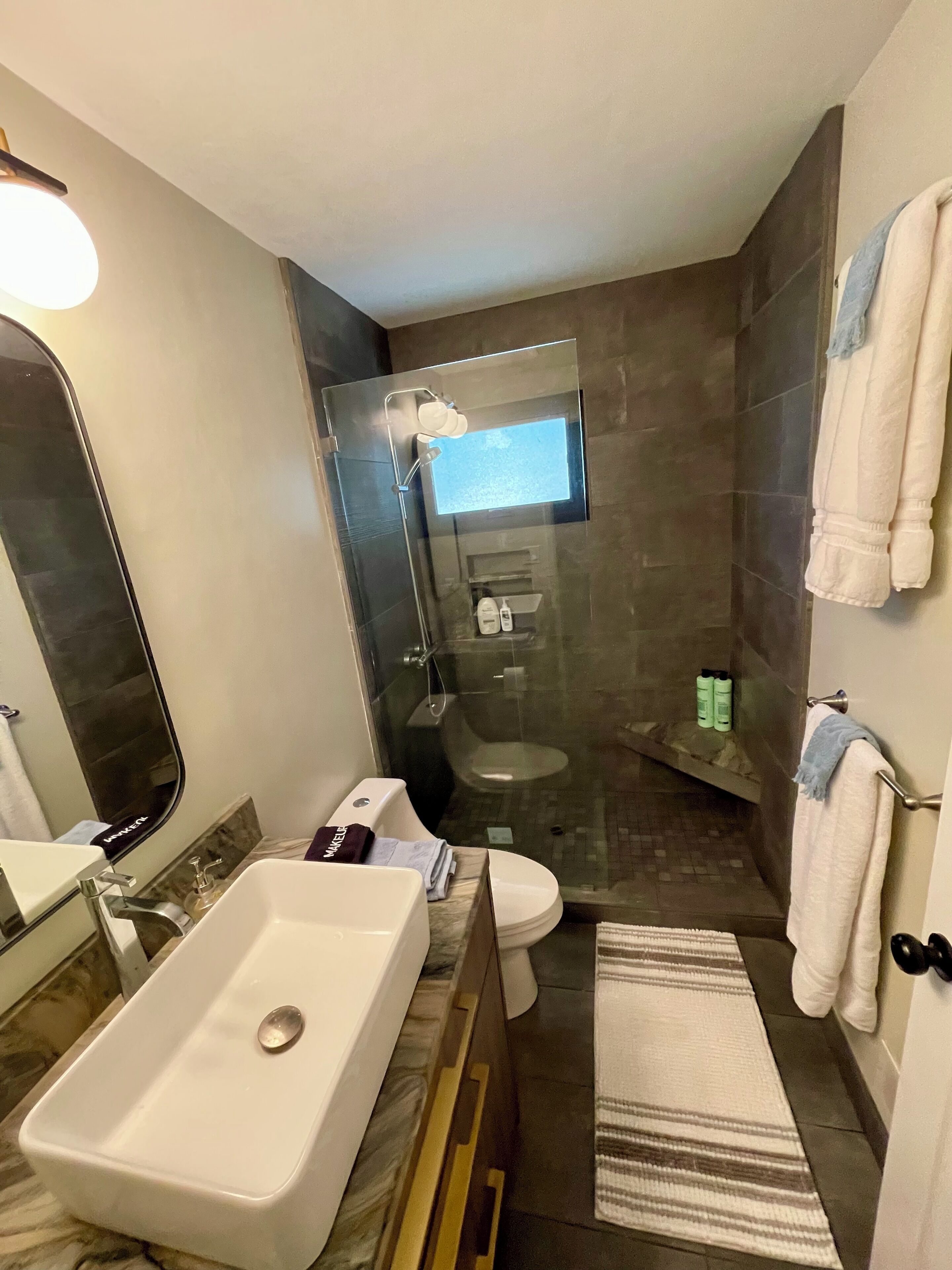 Un baño moderno con ducha de vidrio, un lavamanos blanco, un inodoro y un tocador con espejo.