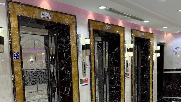 A series of elevators with marble flooring and gold accents, equipped with emergency phones and accessibility signs. - Abd ElQader Naseer Endowment Hotel (Makkah)