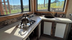 A kitchen with a gas stove, a kettle, and a sink. There are two windows providing a view of a green landscape.