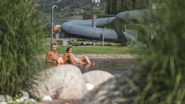 A couple relaxing in a natural pool with a mountainous backdrop and industrial structures. - Cozy apartment farm, with small animals beautiful and quiet location! (Latsch)