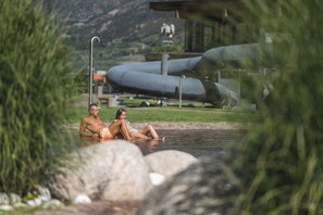 A couple relaxing in a natural pool with a mountainous backdrop and industrial structures. - Cozy apartment farm, with small animals beautiful and quiet location! (Latsch)