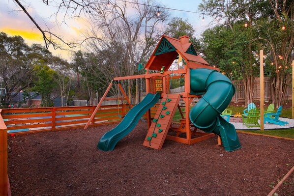 A playground with a slide and climbing area, surrounded by trees and a wooden fence.