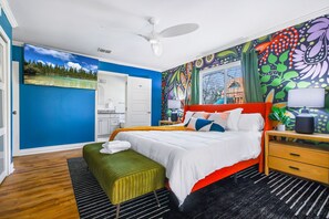 Una camera da letto con un letto, una panca verde, un comodino in legno, una ventola a soffitto e un colorato murale.